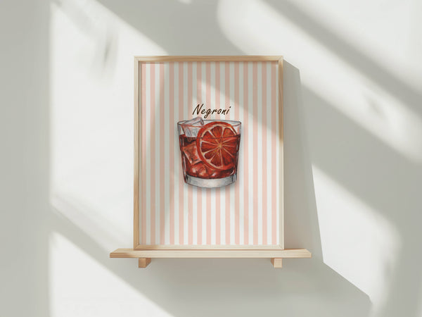 A framed art print featuring a watercolour illustration of a Negroni cocktail in a tumbler glass with ice and an orange slice garnish, set against a background of vertical coral and white stripes. The print is shown in a light wood frame displayed on a wooden shelf against a white wall with soft window shadows.