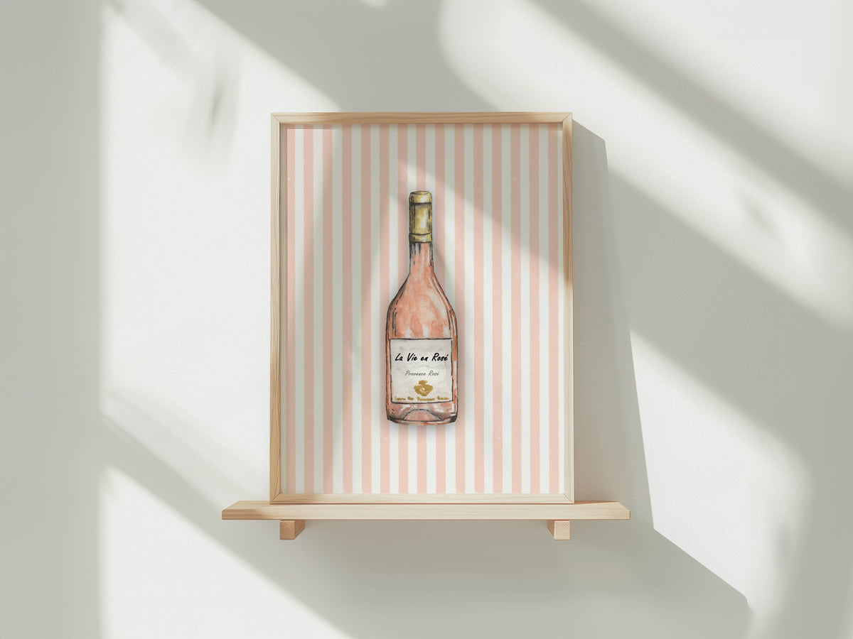 A framed art print featuring a watercolor illustration of a rosé wine bottle against a pink and white striped background, displayed on a light wood floating shelf against a white wall with soft window shadows.