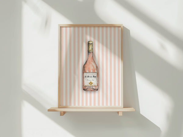 A framed art print featuring a watercolor illustration of a rosé wine bottle against a pink and white striped background, displayed on a light wood floating shelf against a white wall with soft window shadows.