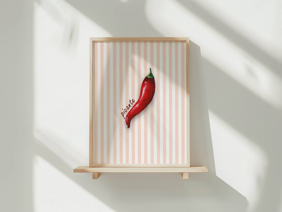 A framed art print featuring a single red chili pepper on a light pink and white striped background, displayed on a light wood shelf against a white wall with soft window shadows.
