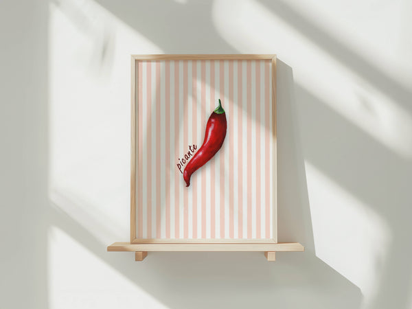 A framed art print featuring a single red chili pepper on a light pink and white striped background, displayed on a light wood shelf against a white wall with soft window shadows.