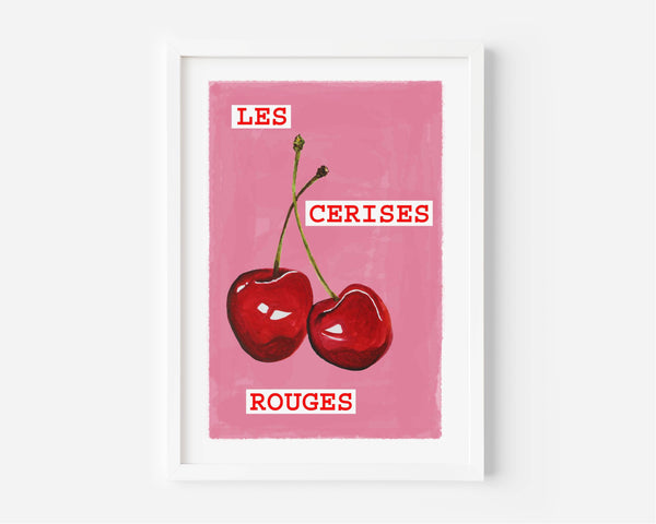 A framed art print featuring two red cherries on a pink background with the French text 'LES CERISES ROUGES' in white lettering, displayed on a light gray wall.