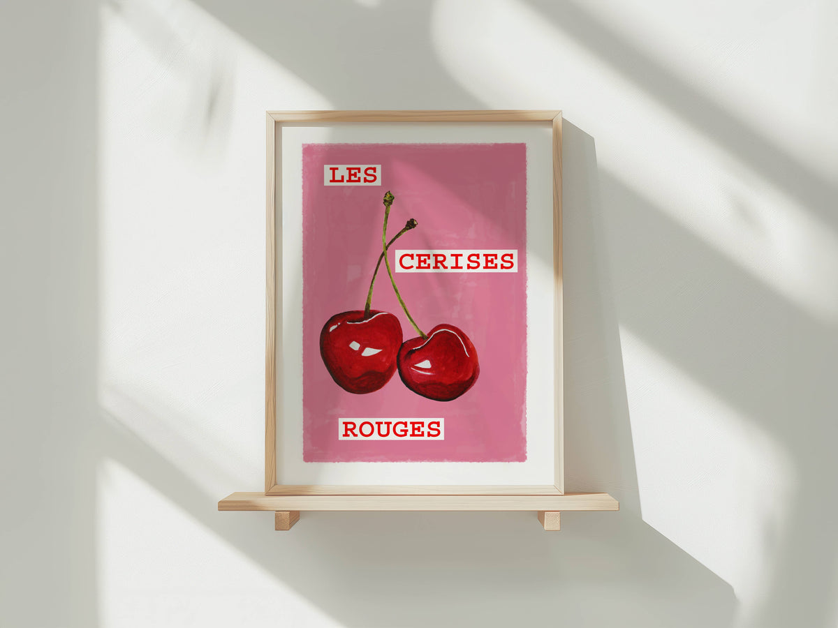 A framed art print featuring a pink background with two red cherries and the French text 'LES CERISES ROUGES', displayed on a light wooden shelf against a white wall with soft diagonal shadows.