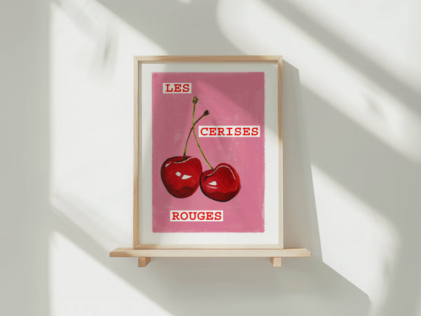 A framed art print featuring a pink background with two red cherries and the French text 'LES CERISES ROUGES', displayed on a light wooden shelf against a white wall with soft diagonal shadows.