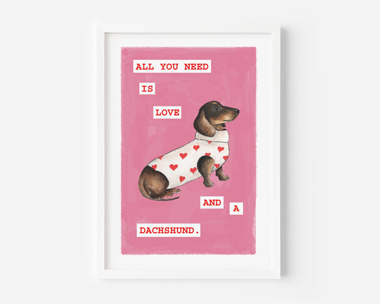 A framed art print featuring a dachshund wearing a white sweater with red hearts on a pink background, with the text 'ALL YOU NEED IS LOVE AND A DACHSHUND.' The print is displayed in a white frame against a light gray wall.