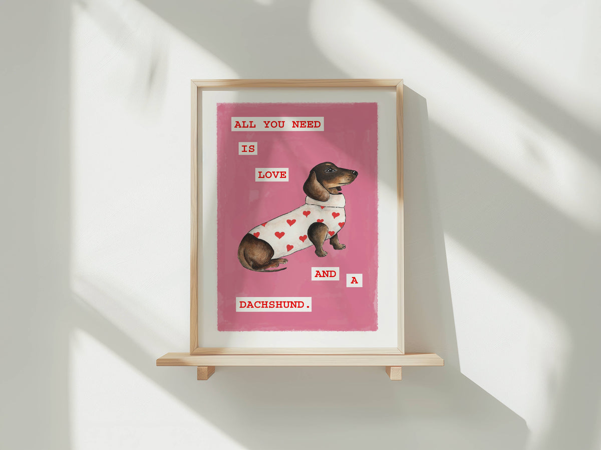 A framed art print featuring a dachshund wearing a white sweater with red hearts on a pink background, displayed on a light wood shelf against a white wall with soft window shadows.