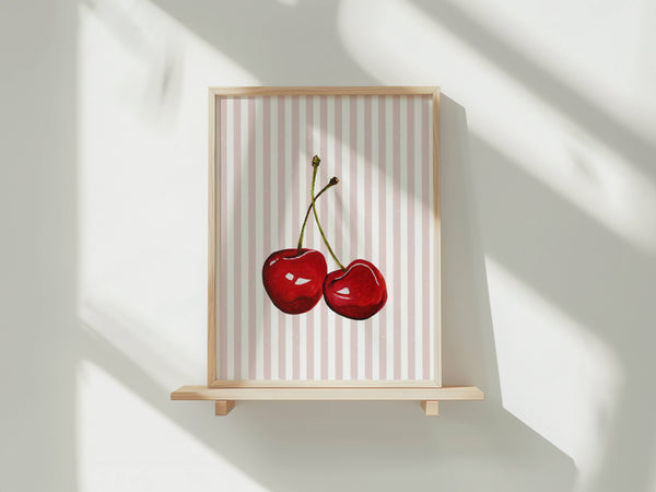 A framed art print featuring two glossy red cherries on a white background with vertical pink stripes, displayed on a light wood shelf against a white wall with soft window shadows.