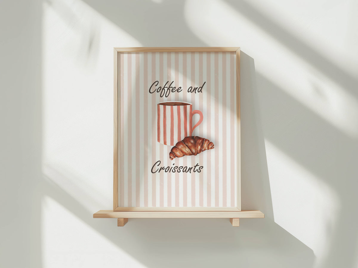 A framed wall art print with a light wood frame and wooden shelf stand, featuring a pink and white striped background with the words 'Coffee and Croissants' and an illustration of a striped coffee mug and croissant. The print is styled on a white wall with soft diagonal shadows.