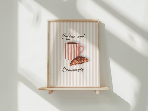 A framed wall art print with a light wood frame and wooden shelf stand, featuring a pink and white striped background with the words 'Coffee and Croissants' and an illustration of a striped coffee mug and croissant. The print is styled on a white wall with soft diagonal shadows.