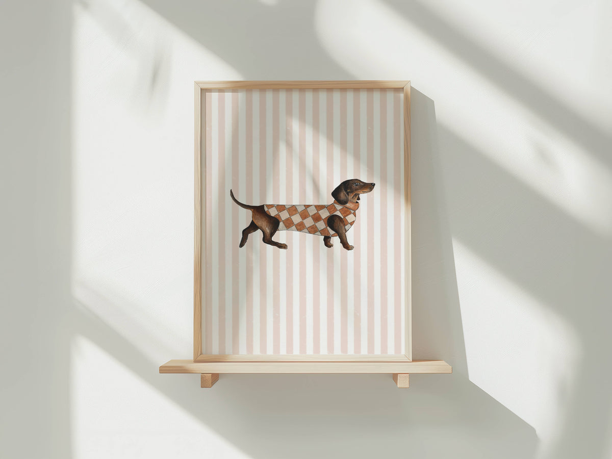 A framed art print featuring a dachshund wearing a brown and beige argyle sweater, displayed on a light wood floating shelf against a white wall with soft window shadows.