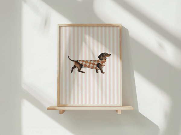 A framed art print featuring a dachshund wearing a brown and beige argyle sweater, displayed on a light wood floating shelf against a white wall with soft window shadows.