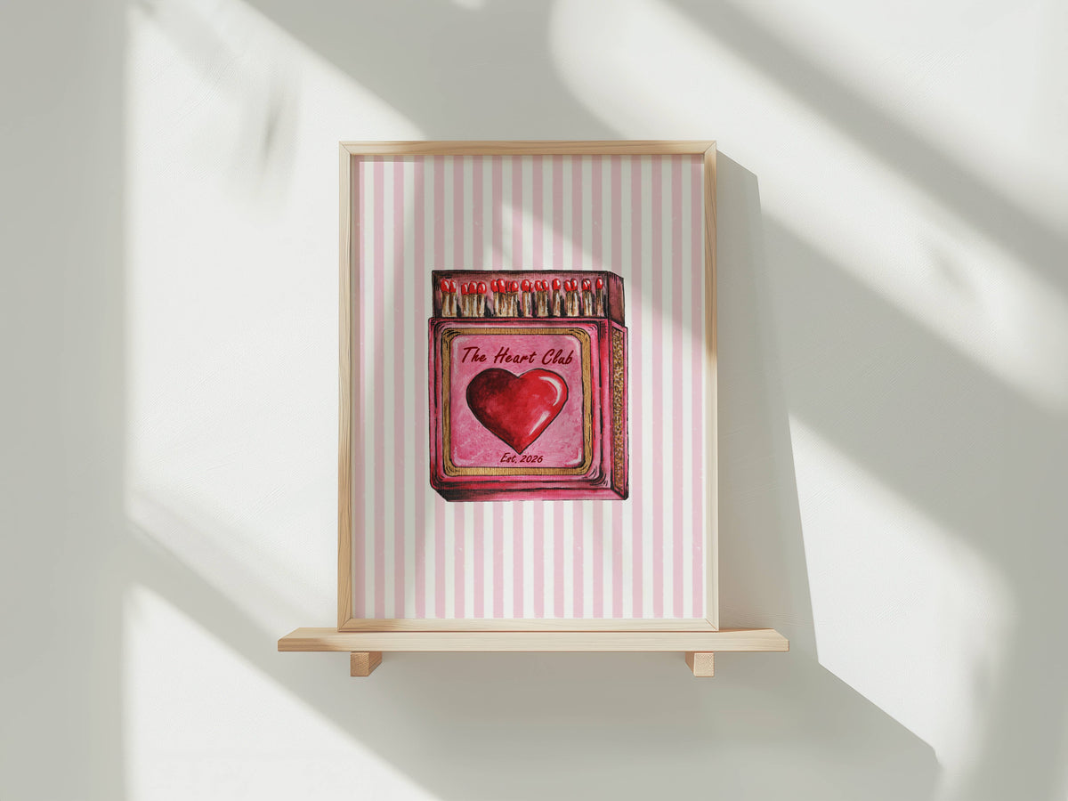 A framed art print featuring a pink and white striped background with a central illustration of a red heart and matchbox design, displayed on a light wooden shelf against a white wall with soft window shadows.