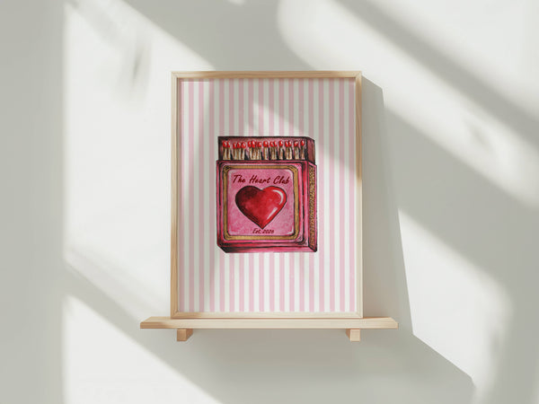 A framed art print featuring a pink and white striped background with a central illustration of a red heart and matchbox design, displayed on a light wooden shelf against a white wall with soft window shadows.