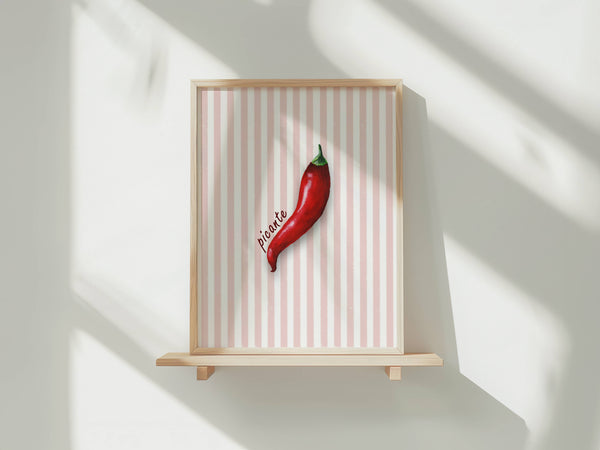 A framed art print featuring a single red chili pepper on a pink and white striped background, displayed on a light wood shelf against a white wall with soft window shadows.