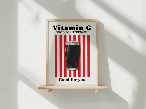 A framed poster on a small wooden stand against a white wall, with soft diagonal shadows. The poster reads 'Vitamin G GOOD FOR STRENGTH' above a glass of dark cola and 'Good for you' below, with red vertical stripes in the background.