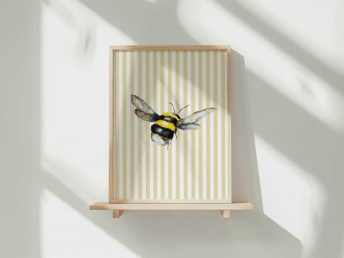 A framed art print featuring a watercolor bumblebee on a cream and beige vertically striped background, displayed on a light wood floating shelf against a white wall with soft window shadows.