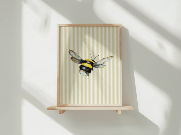 A framed art print featuring a watercolor bumblebee on a cream and beige vertically striped background, displayed on a light wood floating shelf against a white wall with soft window shadows.