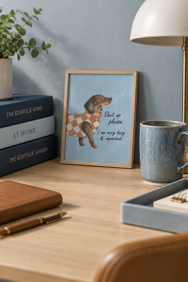 A framed art print featuring a dachshund wearing an argyle sweater with the text 'Shut up please I am very busy & important' displayed on a wooden desk in a styled home office setting.