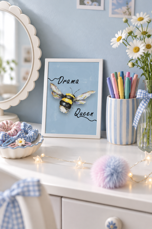 Drama Queen watercolour art print in a small A5 white frame, propped on a girl's dressing table beside a round mirror, hair clips and a vase of daisies, showing a bumblebee on a dusty blue background.