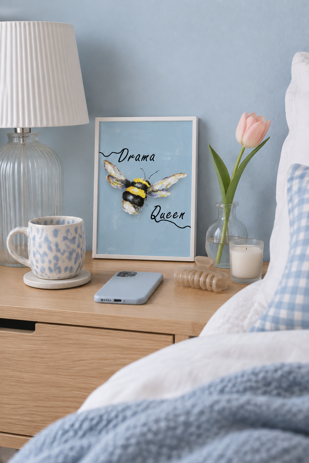 A framed art print featuring a bumblebee with the words 'Drama Queen' in script, styled on a wooden nightstand next to a lamp, a mug, a phone, and a pink tulip in a glass vase.