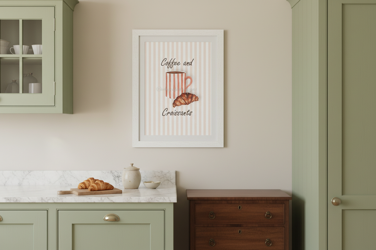 A framed wall art print featuring a striped coffee mug and croissant with the words 'Coffee and Croissant' hangs on a beige wall in a styled kitchen setting with sage green cabinets and a marble countertop.