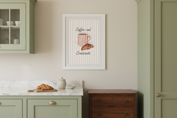 A framed wall art print featuring a striped coffee mug and croissant with the words 'Coffee and Croissant' hangs on a beige wall in a styled kitchen setting with sage green cabinets and a marble countertop.
