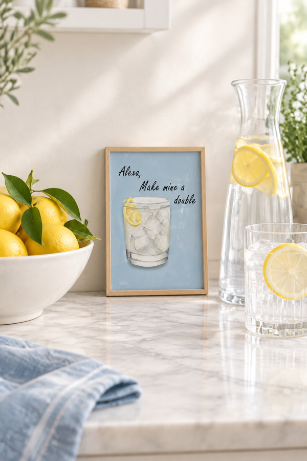 Alexa Make Mine A Double watercolour art print in a small A5 oak frame, propped on a marble kitchen worktop beside a bowl of lemons and a carafe of lemon water, showing a gin and tonic on a soft dusty blue background.