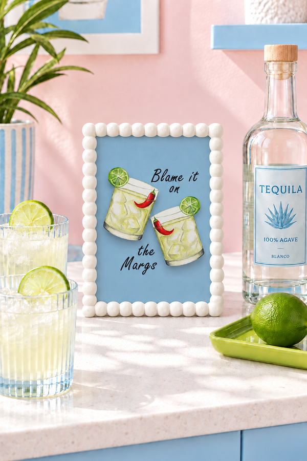 Blame It on the Margs A5 watercolour print in a white bobble frame on a bright home-bar counter, beside cocktail tumblers, a tequila bottle and a lime-green tray against a pale pink wall.