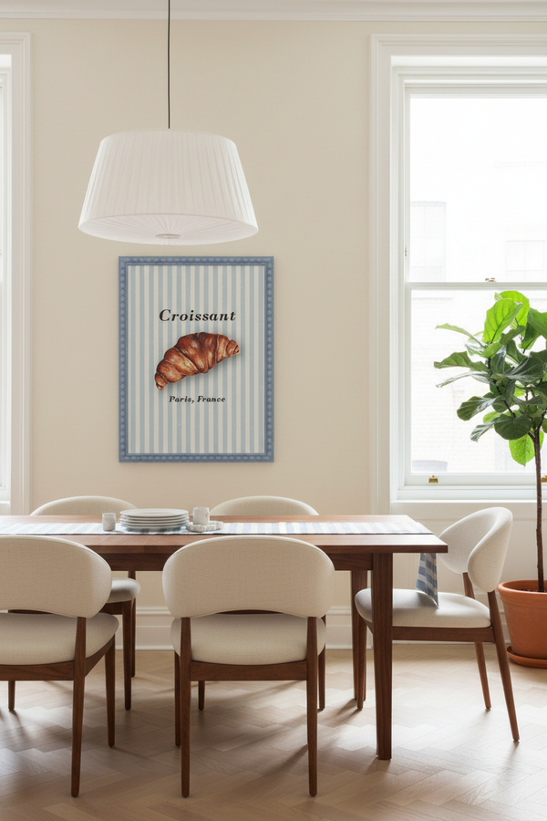 A framed wall art print featuring a croissant on a blue-and-white striped background with the word 'Croissant' and 'Paris, France,' displayed above a dining table in a bright, modern room with chairs, a hanging lamp, and a potted plant.