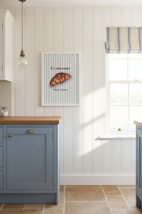 A framed wall art print featuring a croissant on vertical blue-and-white stripes, hung on a white paneled kitchen wall above a blue cabinet in a bright, sunlit kitchen.