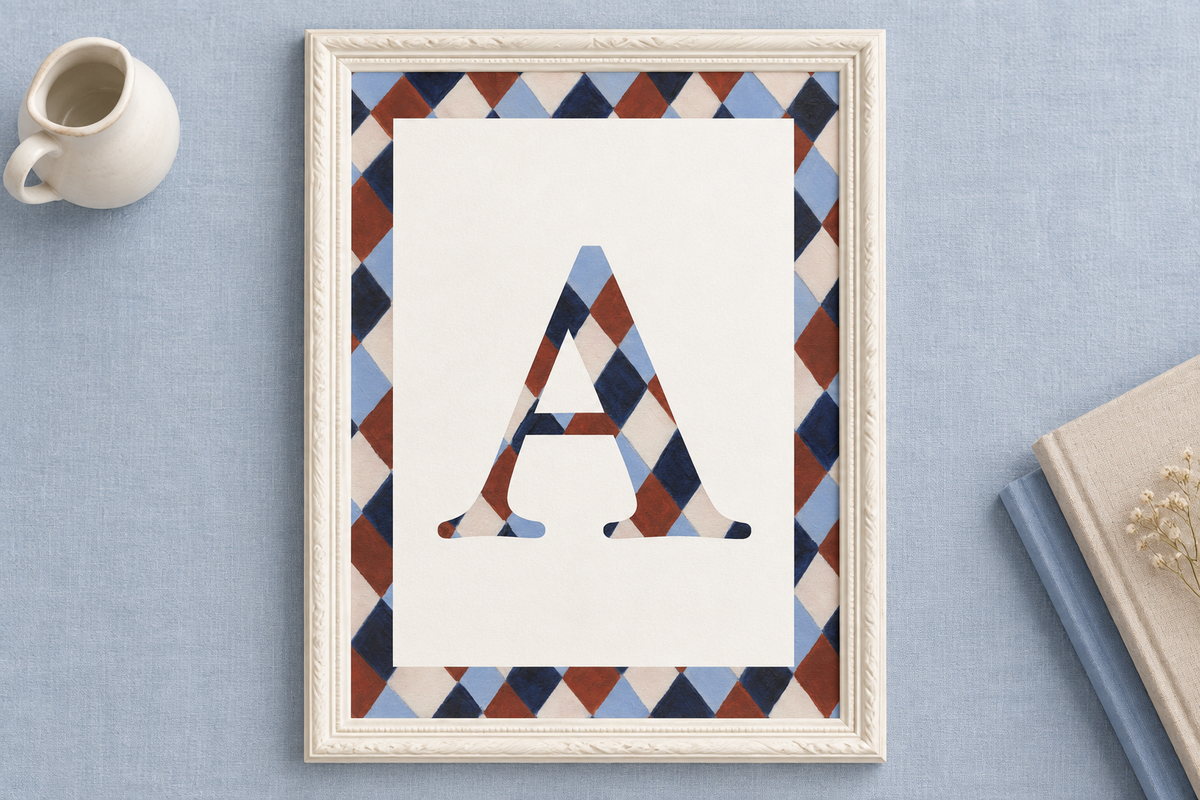 A framed art print featuring a large decorative letter 'A' filled with a blue, brown, and beige harlequin pattern, set against a white background with a matching patterned border. The frame is styled on a light blue textured surface next to a small white ceramic pitcher and a book.