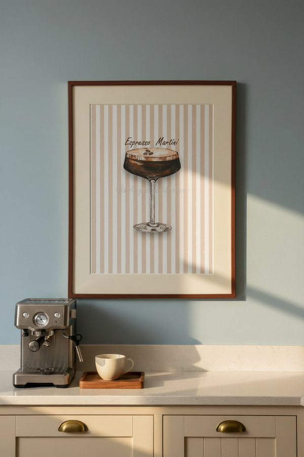 A framed wall art print featuring an espresso martini glass illustration on a cream and beige striped background, hung on a light blue-gray wall above a kitchen counter with a coffee machine and cup.