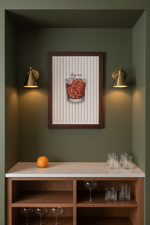 A framed wall art print featuring a cocktail glass with orange slices and the word 'Mojito' on a striped background, displayed in a dark olive green alcove above a wooden bar cabinet with wine glasses and an orange.