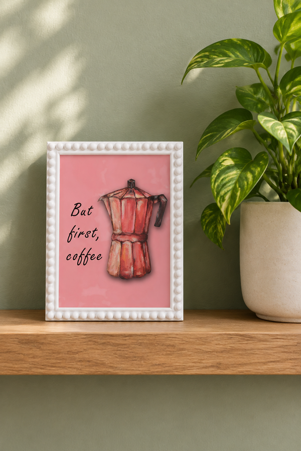 But First Coffee A5 print in a white bobble frame on an oak kitchen shelf beside a leafy plant in a cream pot against a sage wall.