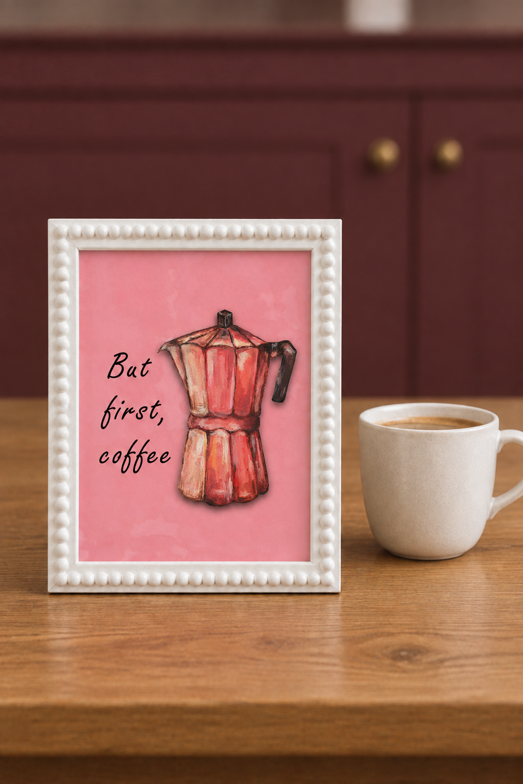 But First Coffee A5 print in a white bobble frame on an oak breakfast table beside an ivory coffee cup, with muted burgundy kitchen cabinetry behind it.