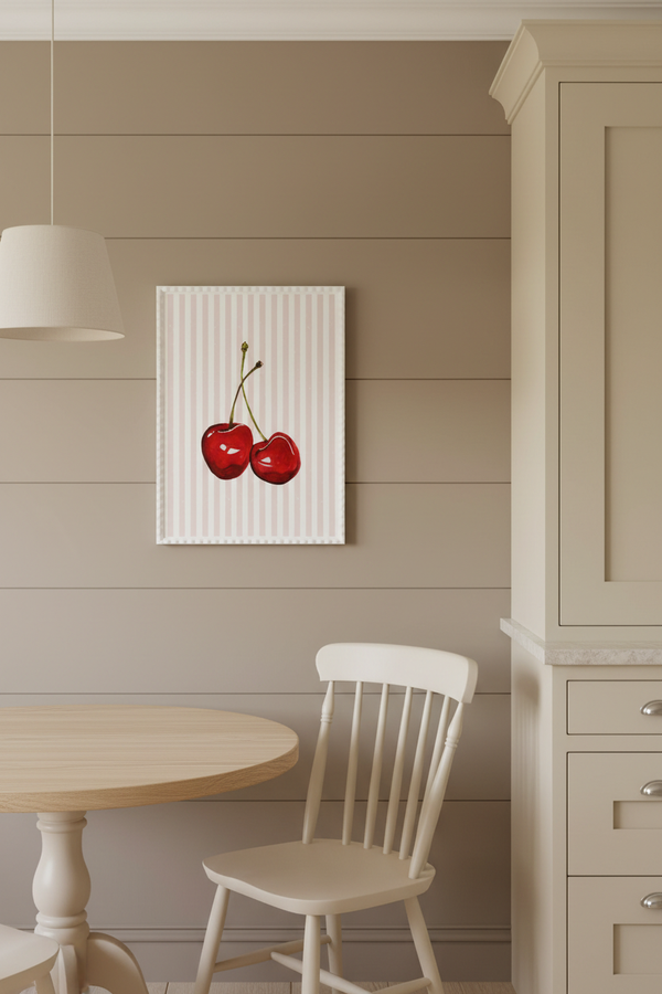A framed wall art print featuring two red cherries on a white striped background, displayed in a dining room setting above a light wood table and white chair.