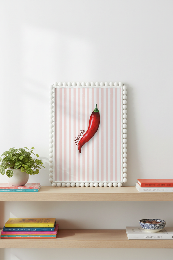 A framed wall art print featuring a single red chili pepper on a white background with pale pink vertical stripes, displayed on a light wood shelf in a styled interior setting with books and a plant.