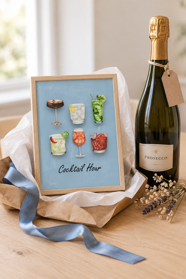 A framed art print titled 'Cocktail Hour' featuring six colorful cocktail illustrations on a light blue background, styled on a wooden table next to a Prosecco bottle and wrapped in tissue paper.