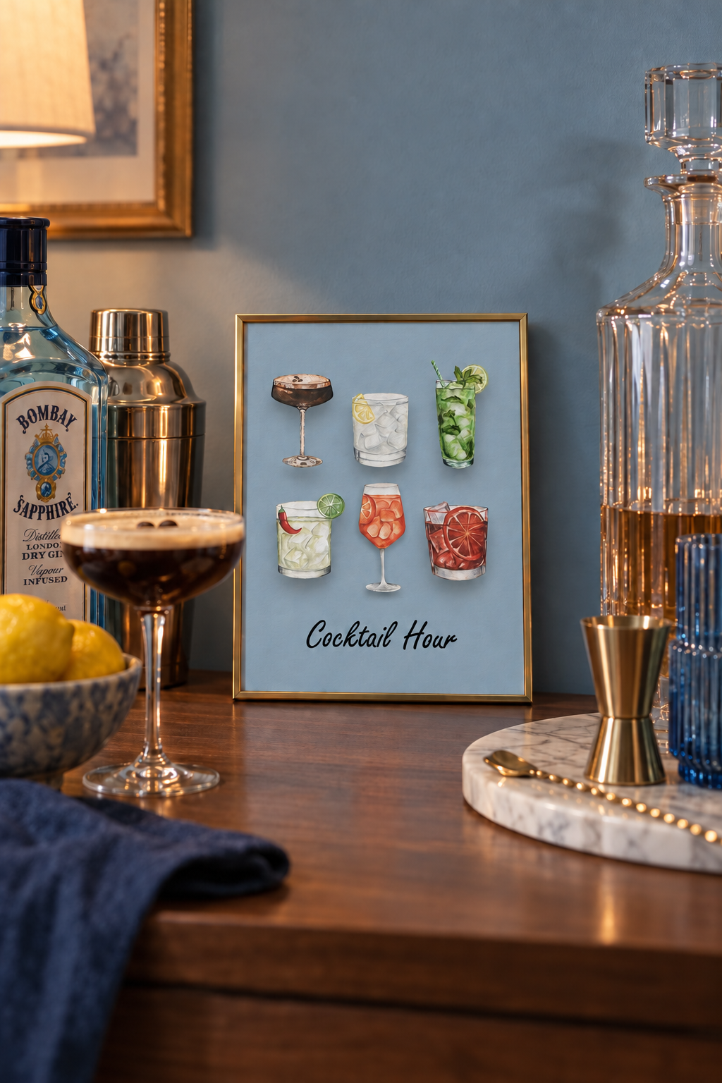 Cocktail Hour watercolour art print in a small A5 brass frame, propped on a home bar shelf beside a gin bottle, cocktail shaker and espresso martini, showing six cocktails on a muted dusty blue background.