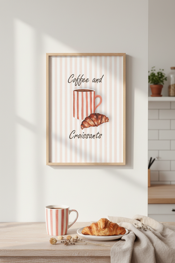 A framed wall art print featuring the words 'Coffee and Croissants' with a striped coffee mug and croissant illustration, displayed above a breakfast table with a matching mug, croissant, and cloth in a bright kitchen setting.