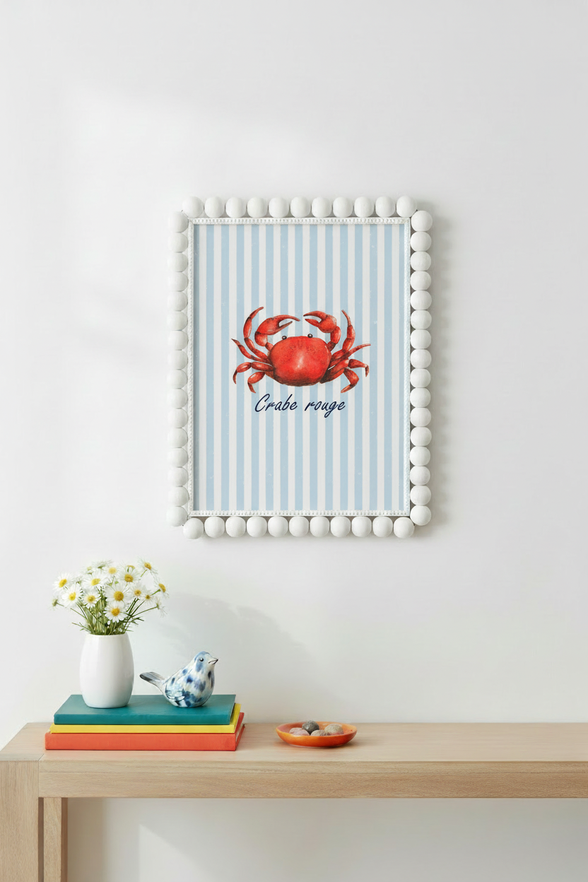 A framed wall art print featuring a red crab on a light blue and white striped background with the text 'Crabe rouge', displayed above a wooden console table with books, a small vase of flowers, and a decorative bird figurine.