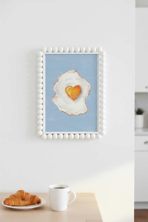 A framed art print featuring a painted fried egg with a heart-shaped yellow yolk on a light blue background, displayed on a white wall above a table with a croissant and a white coffee mug.