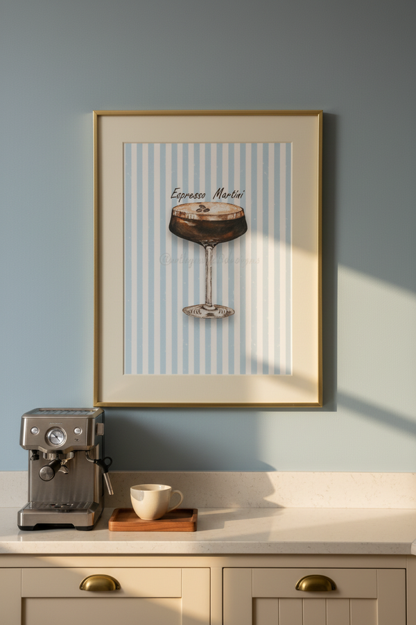 A framed art print featuring an illustration of an espresso martini on a light blue and white striped background, hanging on a light blue wall above a kitchen counter with a coffee machine and cup.