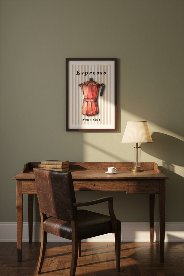 A framed wall art print featuring an espresso moka pot hangs above a wooden desk in a styled room with a leather chair, lamp, books, and a small cup of coffee on a warm olive-green wall.