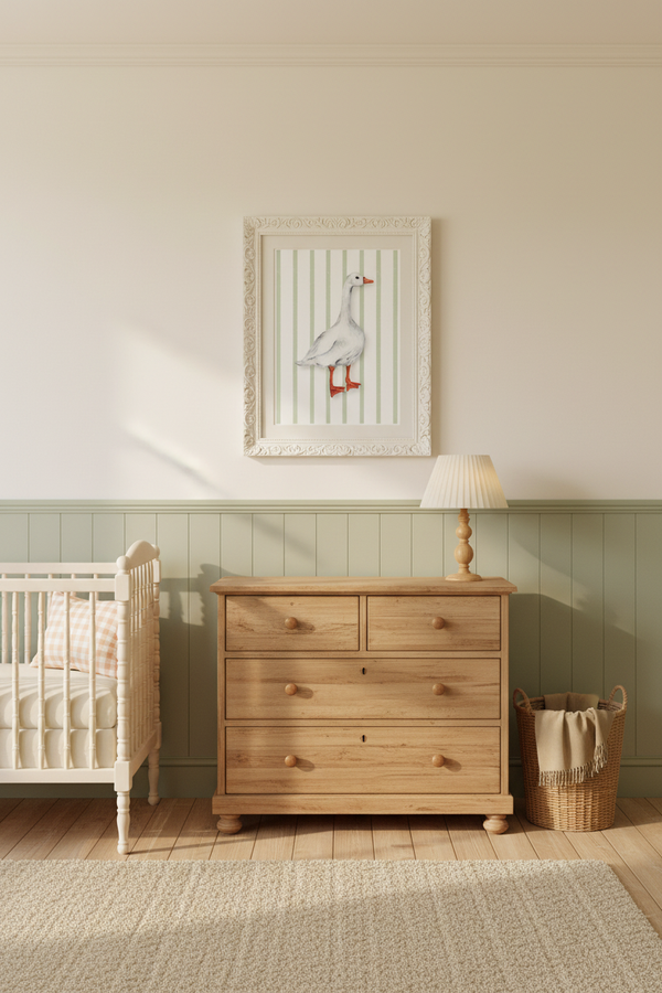 A styled nursery room featuring a light oak wooden five-drawer dresser with round knobs, a matching wooden table with a cream lamp, a white crib, and a framed goose print on the wall.
