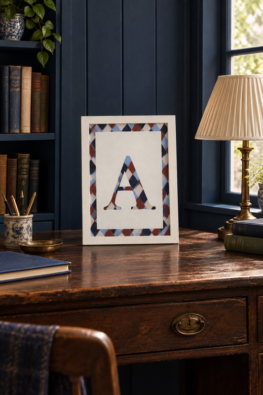 Framed Initial A fine art print in pale blue, navy, warm brown and cream, displayed on an antique writing desk in a cosy inky blue study.