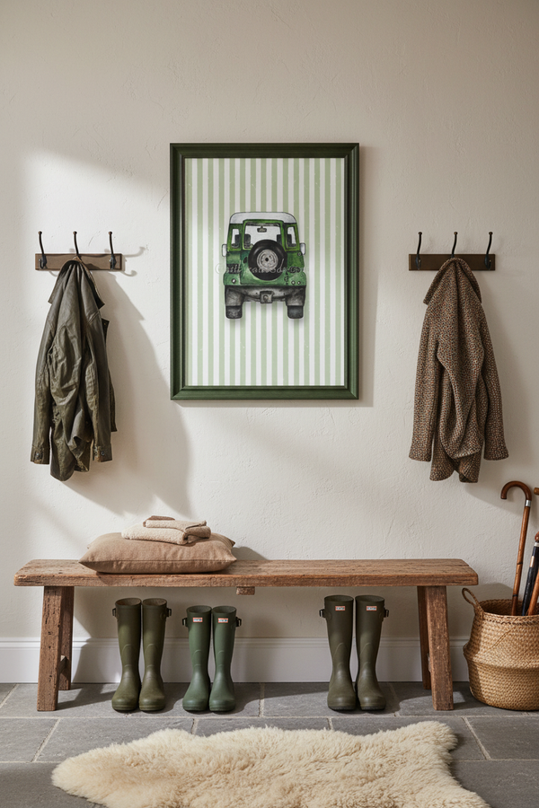 A framed art print featuring a green vintage jeep on a striped background, styled in a rustic entryway above a wooden bench with green rain boots and coats hanging on wall hooks.