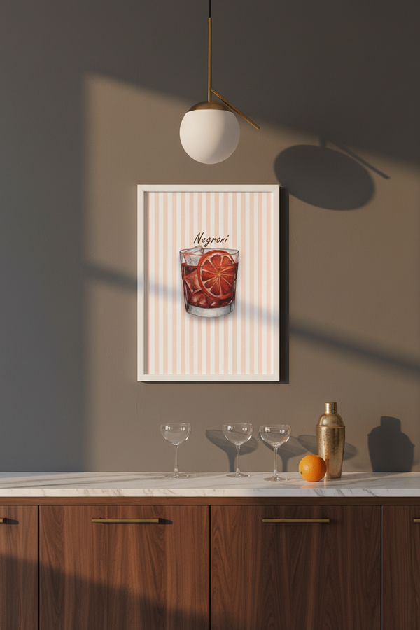 A framed wall art print featuring a cocktail illustration labeled 'Negroni' hangs on a taupe wall above a marble countertop with wine glasses, a shaker, and an orange in a modern kitchen setting.