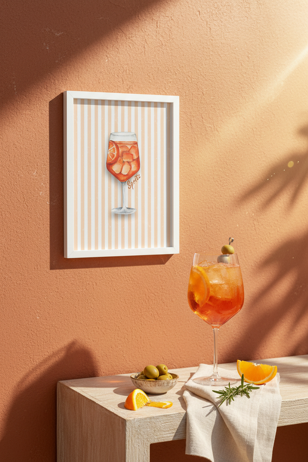 A framed art print featuring a watercolor illustration of an orange cocktail on a striped background, displayed on a warm terracotta wall above a table styled with a matching cocktail, olives, and citrus slices.