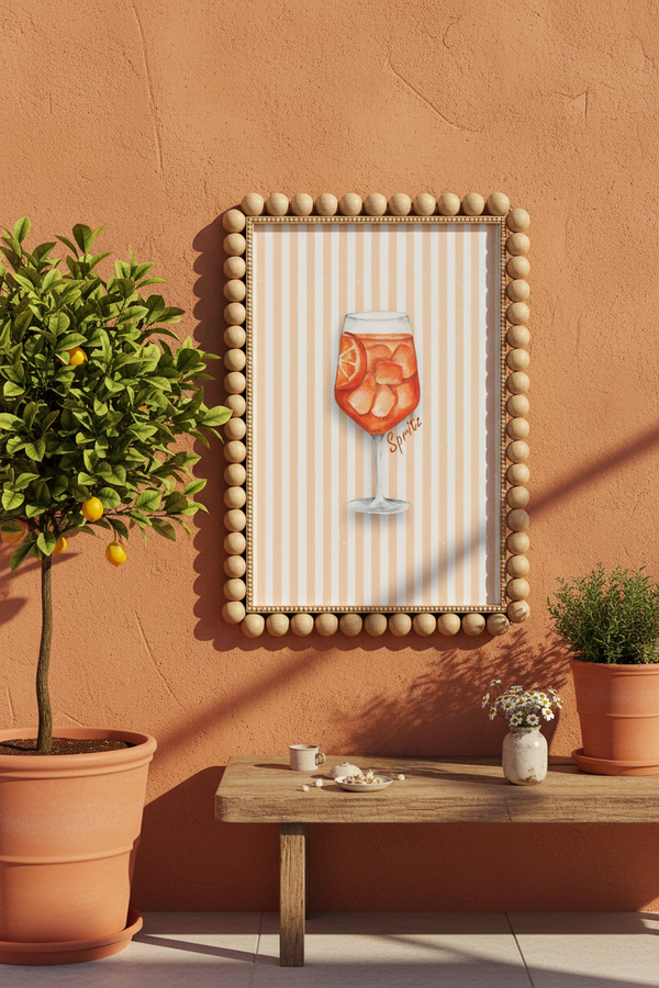 A framed wall art print featuring an orange cocktail on a cream-and-white striped background, displayed on a terracotta wall above a wooden bench with potted plants.